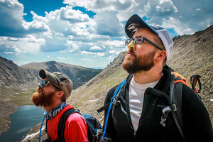 Guy wearing glasses with sunscreen on his nose looking into the sky.  He is wearing a backpack, with a river in the background and mountains around him.